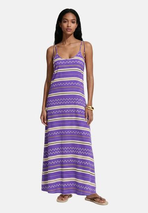 Donna che indossa un lungo vestito viola senza maniche con strisce a zigzag orizzontali beige e bianche, abbinato a sandali e braccialetti dorati.