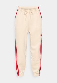 Beige Jogginghose mit roten Seitenstreifen, verjüngten Beinabschlüssen und einem elastischen Bund mit Kordelzug. Mit kleinem Logo an der Seite.