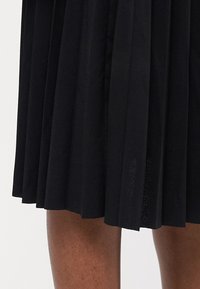 Gonna plissettata nera realizzata in tessuto liscio, caratterizzata da un design di lunghezza media con un leggero luccichio e un aspetto pulito e strutturato.