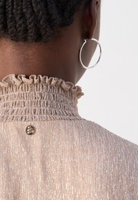 Gros plan sur le cou et l'oreille d'une personne portant une boucle d'oreille argentée en forme d'anneau, vêtue d'un haut doré scintillant à col montant avec un col à volants et un petit bouton doré.