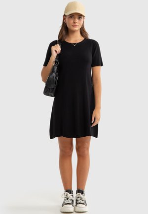 Schwarzes geripptes Strickkleid mit kurzen Ärmeln, einem runden Ausschnitt und einem ausgestellten Saum. Kombiniert mit einer schwarzen Tasche und schwarzen High-Top-Sneakern.