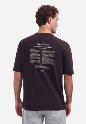 Man met krullend haar die van zich af kijkt, draagt een zwart T-shirt met witte bedrukte tekst getiteld "Sahara Simplicity" op de bovenrug.