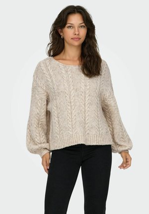 Maglione in maglia a treccia di colore crema chiaro con un ampio scollo e maniche a palloncino, con orlo a coste. Indossato con pantaloni aderenti neri.