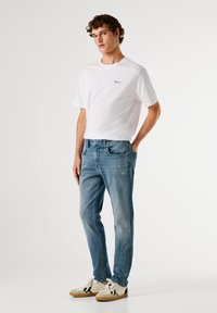 Camiseta blanca de algodón con logo rojo, jeans de mezclilla azul claro desgastados y zapatillas crema con suela marrón, acentos en negro y azul.