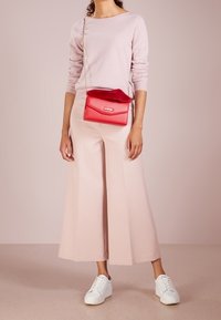 Světle růžový svetr, široké světlé růžové kalhoty, červená kabelka s kožešinovým akcentem a bílé tenisky. Hladké textury a čisté linie.
