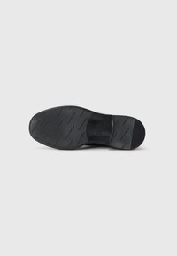 Semelle de chaussure noire avec surface texturée, présentant des rainures horizontales pour une adhérence optimale et un bout arrondi. Design minimaliste avec un logo embossé.