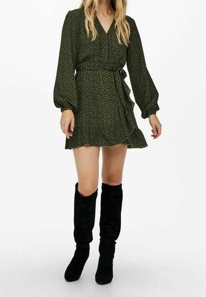 Frau trägt ein dunkelgrünes Wickelkleid mit langen Ärmeln und schwarzen kniehohen Stiefeln, steht vor einem schlichten weißen Hintergrund.