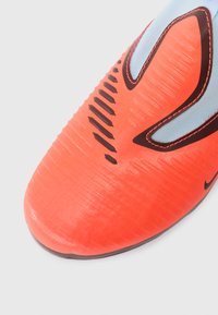 Κοντινό πλάνο από το άκρο μιας υφασμάτινης ποδοσφαιρικής πατούσας σε κόκκινο και μαύρο χρώμα, με ορατές ραφές και ένα μικρό μαύρο λογότυπο σε λευκό φόντο.