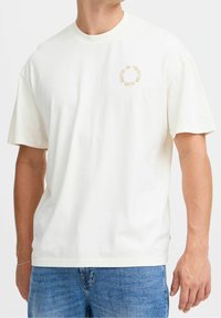 T-shirt en coton blanc avec un col rond et des manches courtes, présentant un design graphique circulaire en texte beige sur la poitrine.