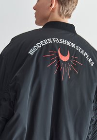 Czarna kurtka bomberka o kroju cropped, z elastycznymi mankietami oraz czerwonym motywem graficznym z tekstem i półksiężycem na plecach.