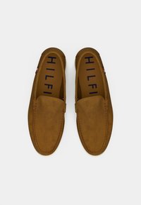 Mocasines de ante marrón con una textura suave y detalles de costura. El interior presenta "HILFIGER" en letras azules. Suela de goma ligera.