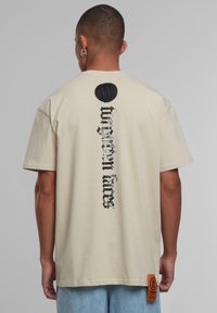 Krémové tričko s krátkými rukávy, s vertikálním černým grafickým textem a kulatou nášivkou u výstřihu. Štítek na dolní straně.