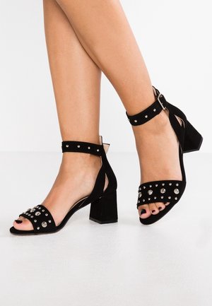 Sandali aperti neri in suede con borchie in argento, tacco blocco e cinturino alla caviglia con fibbia per una vestibilità sicura. Texture liscia su tutta la superficie.