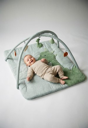 Lächelndes Baby in beigem Outfit, liegend auf einer grünen Dinosaurier-Spielmatte mit weichen Bögen und hängenden Plüschspielzeugen.