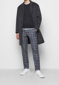 Mörkgrå ullkappa över en svart tröja. Ljusgrå och blå rutiga byxor i kombination med vita sneakers. Enkla, rena linjer och skräddarsydd passform.