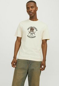 Krém színű pamut póló, középen egy kalapos koponyát ábrázoló grafikával, körülötte a "JACK&JONES WORLDWIDE" felirattal és villámcsapásokkal.