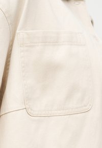 Nærbilde av en offwhite jakkelomme i stoff med synlige sømmer og en knapp nær kragen.