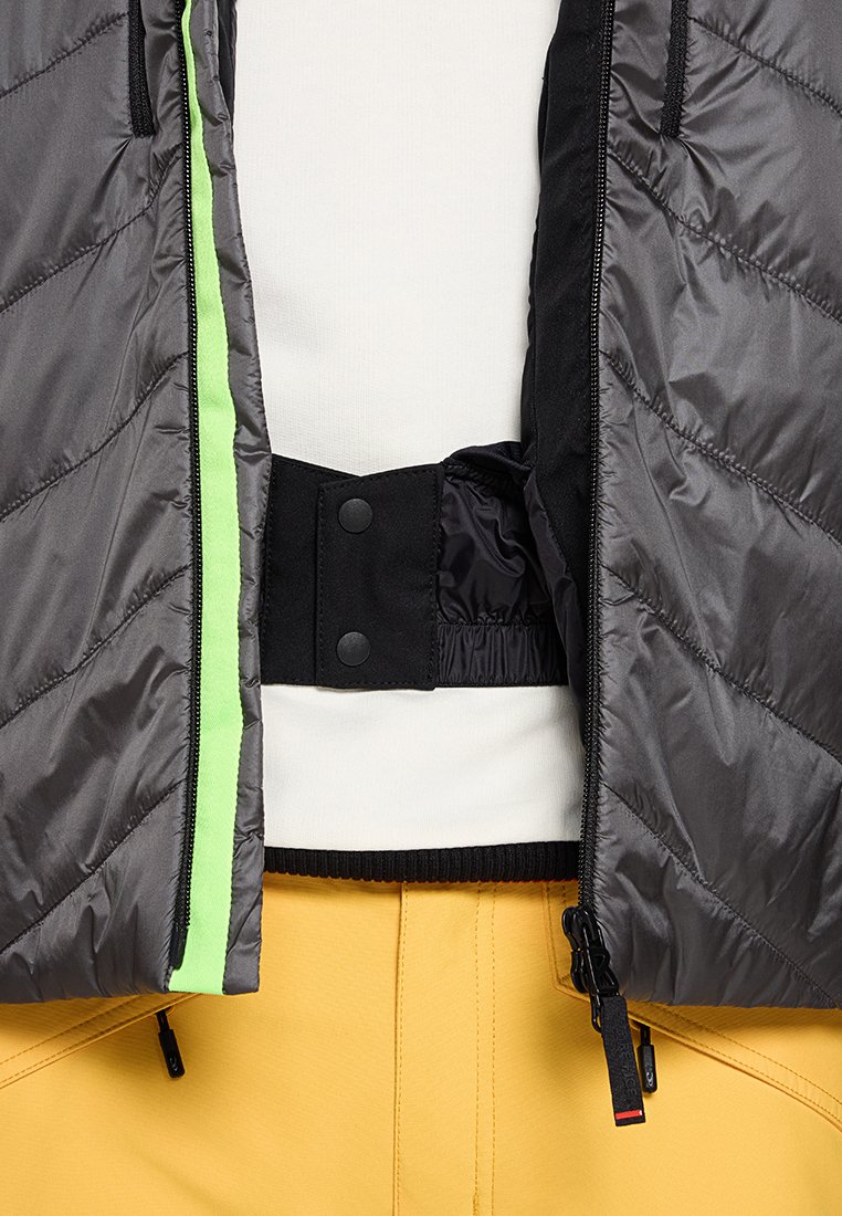 Grijze pufferjack met diagonale quilt, groene ritssluiting, zwarte elastische band en drukknoopsluiting. Draagt over een witte top en gele broek.