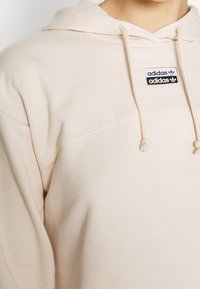 Ljusbeige hoodie tillverkad av mjukt tyg, med en dragsko i huvan och en svart Adidas logopatch på bröstet. Sömnadsdetaljer är synliga.