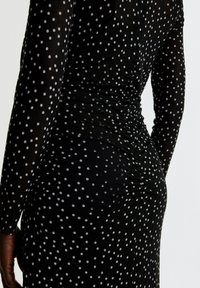 Schwarzes Kleid mit langen Ärmeln, das mit weißen Punkten versehen ist. Der Stoff ist am Rücken gerafft und betont eine taillierte Silhouette.