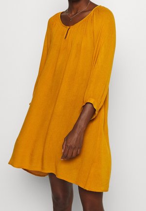 Femme portant une robe jaune moutarde ample à manches longues, longueur genou, avec un petit détail de bouton au niveau de l'ouverture en forme de goutte.