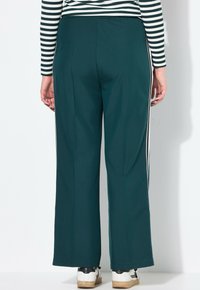 Persoon met donkergroene wijde broek met witte zijstrepen, een zwart-wit gestreepte lange mouw top, en witte sneakers, met de rug naar de kijker gekeerd.