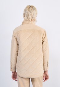Beige kordfløyels skjortejakke med et quiltet mønster, som har krage og knappede mansjetter, og viser en teksturert og ribbet overflate.