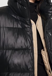 Svart puffjacka med en blänkande yta, hög krage och dragkedja, över en beige innerjacka med synliga fickor och sömmar.