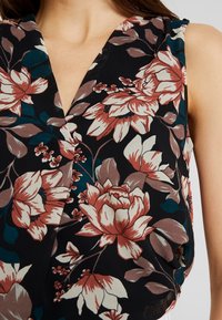 Blusa nera floreale con grandi fiori bianchi e ruggine, accompagnati da foglie verdi. Design a V e tessuto morbido e fluido. Senza hardware visibile.