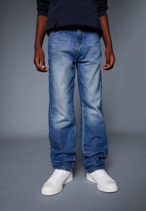 Person iført løstsiddende blå jeans, hvide sneakers og en mørk langærmet top, stående foran en ensfarvet grå baggrund.