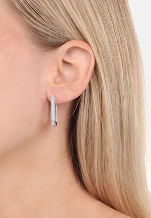 Orecchino a cerchio in argento con superficie texture, impreziosito da piccole pietre preziose trasparenti, tenuto da una chiusura elegante, indossato all'orecchio di un modello.