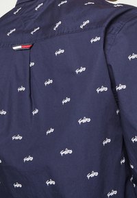 Granatowa bawełniana koszula z białym nadrukiem logo, klasycznym kołnierzykiem i detalem w postaci zakładki z tyłu. Prosty design z gładką teksturą.