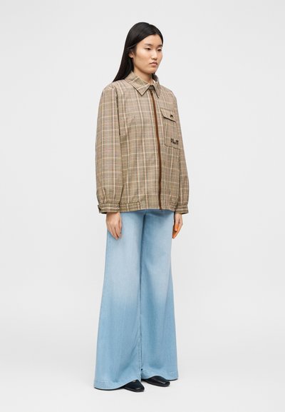 Καφέ καρό φαρδιά ζακέτα με τσέπη στο στήθος και λεπτομέρεια φερμουάρ, συνδυασμένη με ανοιχτόχρωμο μπλε παντελόνι με φαρδιά πόδια και μαύρα παπούτσια.