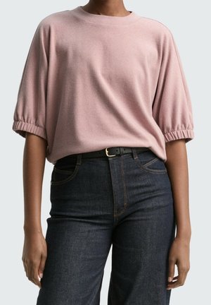 Frau trägt ein lockeres, hellrosa kurzärmeliges Oberteil mit gerüschten Ärmeln, das in eine dunkelblaue, hoch taillierte Jeans mit schwarzem Gürtel gesteckt ist.