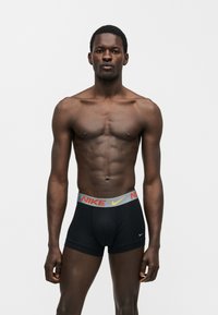 Homme athlétique debout, face à l'avant, portant un boxer noir Nike avec une ceinture grise et un logo orange sur un fond blanc uni.