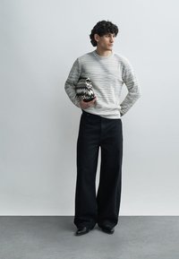 Szürke-fehér csíkos pulóver, fekete bő szárú nadrág, fekete-fehér cikk-cakk mintás táskát tartva, világos háttér előtt állva.