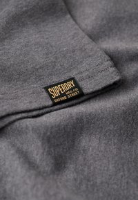 Grå stof med en blød tekstur, der har et "Superdry" mærke i sort og guld på den nederste kant.