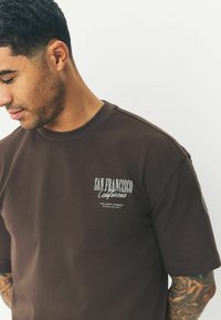 Brun bomulds T-shirt med korte ærmer, med hvid tekst der står "San Francisco California" og "Folsom Street Mission District."