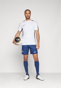 Witte Nike sportshirt met zwarte accenten, blauwe short met een logo, blauwe sokken en een zwart-witte voetbal.