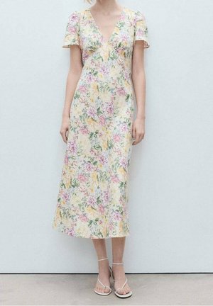 Kvinde iført en pastel-farvet blomsterprint midi-kjole med korte ærmer og V-hals, kombineret med hvide, hælede sandaler med stropper, stående foran en ensfarvet baggrund.