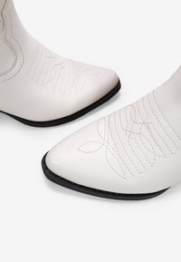 Botas de tobillo de cuero blanco con puntas afiladas, que presentan patrones de costura sutiles y una suela negra contrastante. Textura suave con acentos mínimos.