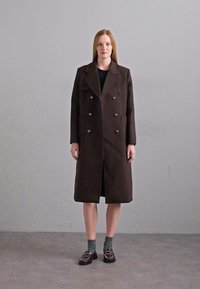 Manteau marron croisé avec de grands revers, poches latérales et une silhouette droite. Porté par-dessus un haut noir, associé à des chaussettes grises et des mocassins.