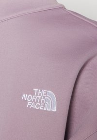 Světle fialová polo košile s vyšitým bílým logem "The North Face" na levé straně. Hladká textilie a jednoduchý design.