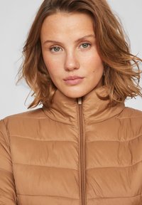 Kahvatugevusega jakk kõrge kraega, millel on õmbluse detailid ja eesmine tõmblukk. Sile tekstuur ja liibuv disain.