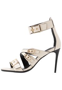 Sandal de tacón alto beige con diseño de tiras, que presenta tres hebillas doradas y una suela negra. Textura de cuero suave y puntera afilada.