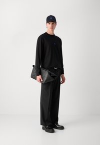 Zwarte langearmige shirt, bijpassende broek en pet. De figuur houdt een gestructureerde zwarte clutch met geometrische accenten vast. Zwarte schoenen maken de look compleet.