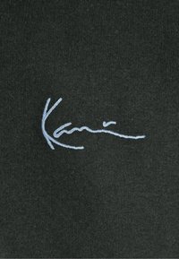 Mørkegrønt stof med et lyseblåt broderet signaturdesign, glat tekstur med en tætsnittet vævning, centreret på tøjet.