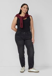 Schwarzes ärmelloses Oberteil, dunkelgraue Jeans mit vorderen Taschen und cremefarbene Sneakers mit olivgrünen Akzenten. Weinroter Pullover lässig um die Schultern gebunden.