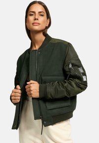 Zelená bomber bunda s hladkou textúrou, so zipsovým vreckom na ľavom rukáve, rebrovanými manžetami a dvoma prednými vreckami. Obsahuje kontrastné látkové panely.