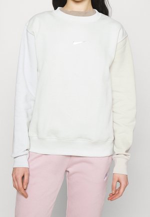 Personne portant un sweat-shirt blanc Nike à manches longues et un pantalon de survêtement rose Nike, debout devant un fond uni.
